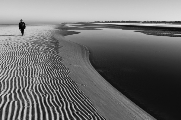 das schwarzweißfoto im querformat zeigt eine einsame person, die an einem sandstrand entlanggeht, der von einem ruhigen wasserlauf begrenzt wird.

ein warm gekleidete person mit rucksack ist in einiger entfernung an der linken bildkante zu sehen.
der sand ist von deutlichen, wellenförmigen mustern geprägt, die durch die wirkung der gezeiten entstanden sind. der strand ist breit und erstreckt sich bis zum horizont, wo eine flache küstenlinie mit dünen zu sehen ist. links das offene mehr; diese küstenlinie geht nach rechts über in die feine linie der dünenhügels.
ein ruhiger wasserlauf schlängelt sich nach hinten hoch entlang der kante zum unteren strand, an dem die person zu sehen ist.
das wasser ist ruhig mit einer glatten oberfläche, es spiegelt das helle grau des himmels wieder, das nach unten dunkler wird.

das bild ist kontrastreich. das direkte licht der sonne sorgt für eine starke betonung der linien und texturen. die stimmung ist melancholisch und einsam, was durch die einsamkeit der person und die weite der landschaft verstärkt wird.