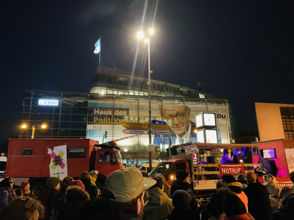 A nighttime protest scene in front of a building with a large advertisement featuring a person. Two red trucks are visible, one displaying a sign that reads "AfD Verbot Jetzt" and another with a "Notruf" sign. A crowd of people