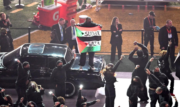 Un danseur debout sur la voiture noire de la scène, habillé en noir et un bandana sur le visage, brandi un drapeau du Soudan et de la Palestine avec  : 🖤SUDAN ✊🏿GAZA inscrit dessus