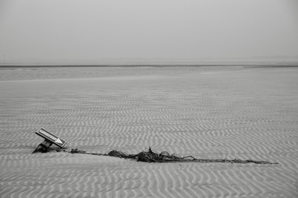 das schwarzweißbild im querformat zeigt einen sandstrand an einem trüben tag.
im vordergrund liegt links ein umgefallenes und halb im sand versunkenes hinweisschild mit der aufschrift "rundgang" und einem weißen richtungspfeil.der holzbalken, an dem das hinweisschild hängt, ist umwickelt mit sich auflösenden tauen und schnüren. die schnüre verlaufen vom schild links bis nach rechts an den bildrand in fast waagerechter linie.
der hintergrund zeigt einen weitläufigen, flachen strand mit wellenförmigem muster und dem flachen meer dahinter. in der ferne verschwindet der horizont im trüben und nebligen dunst.