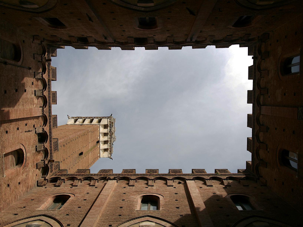 Das Bild zeigt den Torre del Mangia in Siena, Italien. Das Bild wurde vom inneren des Palazzo Publico aufgenommen. Der Blick geht senkrecht nach oben, so dass der Blick gen Himmel von den Dächern im Form eines Rechteck gesäumt wird. Der Turm steht auf einer Ecke dieses Rechtecks.