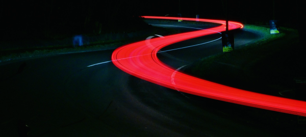 In dem überwiegend schwarzen Bild ist in einer S-Kurve einer geteerten Straße die dünne weiße Linie des Mittelstreifens sowie ein breiter leuchtend roter Streifen des durchgehenden Rücklichts eines Elektroautos zu sehen, der in einer Langzeitaufnahme dem eleganten Schwung der Kurve folgt und über der Straße zu schweben scheint.