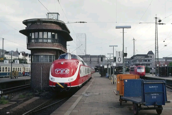 Alt-Text: Ein klassischer Trans-Europ-Express (TEE) in roter Lackierung mit aerodynamischer Front steht an einem Bahnsteig. Links daneben befindet sich ein historischer Stellwerksturm mit runder Fensterfront und einer Uhr auf dem Dach. Im Hintergrund sind moderne Gebäude sowie ein weiterer roter Zug zu sehen. Der Bahnsteig ist mit Gepäckwagen und Schildern mit Gleisnummern versehen. Die Szene vermittelt eine Mischung aus Nostalgie und moderner Bahninfrastruktur.

