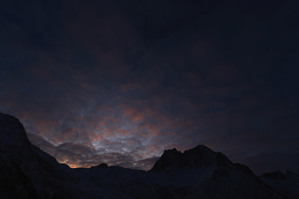 Just a hint of light over the horizon, the profile of a mountain stands in the darkness while fluffy clouds high over head are painted peach and pink.