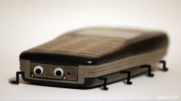 A photo of an old mobile phone against a white backdrop. It is on its back, base towards the camera, and the photo is taken low down with a very tight focus. The light was low so it's a little grainy. Googly eyes have been added over two sockets at the bottom, and the edge of the panel reads as a mouth. I've accentuated it very slightly and added a couple of upturned fangs. The body has little legs added, 4 aside. 