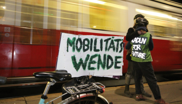 Zwei Radelnde mit der Aufschrift "Radeln for Future" und einem Demo-Schild "Mobilitätswende" vor einer vorbeifahrenden Straßenbahn.