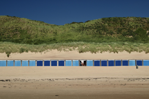 das bild im querformat zeigt oben im bild eine grün bewachsene düne im hintergrund. im bild das untere vom mittleren drittel abtrennend sind als horizontale linie strandhütten zu sehen, die zur lagerung von liegestühlen und anderen accessoires benutzt werden. die hütten sind in hellblau und in dunkelblau gestrichen.
die türen einer diesen hütten in der bildmitte sind geöffnet, und eine person in badehose nimmt auf einem campingstuhl ein sonnenbad.
in der unteren bildhälfte ist der sand zu sehen. 
im hintergrund hinter den dünen ist dunkles sattes blau des wolkenlosen himmels zu sehen.
insgesamt ein sehr gesättigtes bild mit kräftigen farben. die farben blau, grün und gelb sind dominant.