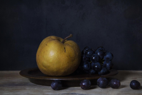 A dark still life photo of a bronze Nashi pear and blue grapes placed on a brown ceramic plate on a wooden board. Some individual grapes are randomly scattered in front of and on the side of the plate. The background is dark slate gray. The whole scene has an old-fashioned, painterly feel.