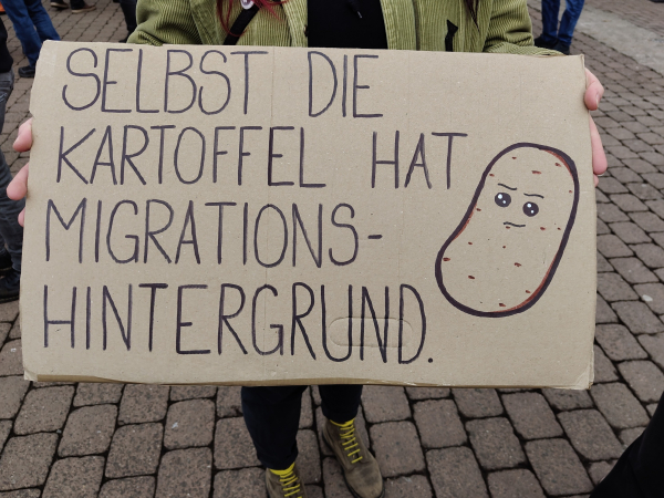 Selbstgemaltes Plakat. Rechts ist eine Kartoffel mit Gesicht abgebildet. Daneben steht der Text: "Selbst die Kartoffel hat Migrationshintergrund."