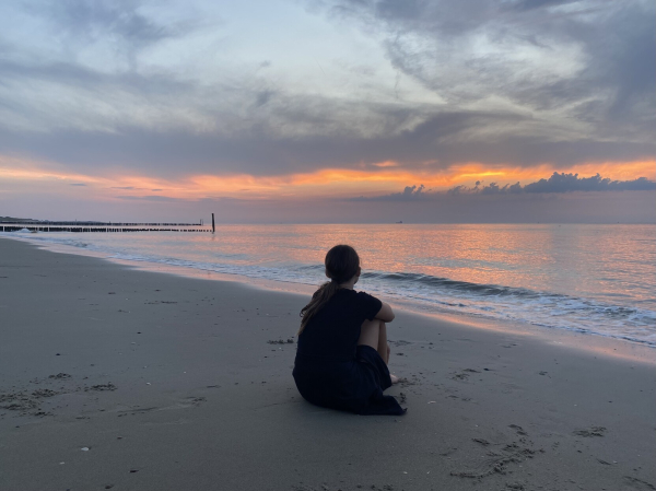 Sonnenuntergang, Vordergrund Strand ein Mädchen sitzt mit dem Rücken zur Fotografin, relativ glatte Wasseroberfläche 