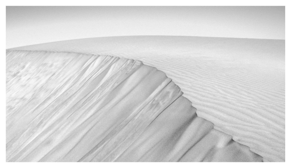 This rectangular black-and-white photograph, framed in white, captures the elegant curves and textures of a sand dune. The composition emphasizes the contrast between the smooth, wind-sculpted ridges at the top and the sharply defined, cascading layers of sand on the slope below. Gentle ripples stretch across the surface. The monochrome palette enhances the abstract quality of the scene, making the fine details of the shifting sand appear almost like flowing fabric. The vast, empty background further adds to the sense of isolation and timelessness.