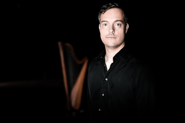 Harpist standing in front of a harp that is fading into a black background. 