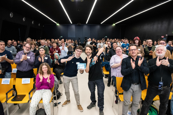 Matthias and Raphael at SFSCON 2024 in the middle of a crowd after asking all people in the audience to stand up if are using Vim, or have used Vim in the past. Matthias showing the VI M sign with his fingers - CC-BY-SA 4.0 NOI