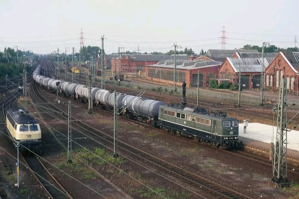 Alt-Text: Eine dunkelgrüne Elektrolokomotive der Baureihe 151 zieht einen langen Kesselwagenzug durch einen weitläufigen Rangierbahnhof mit alten Backsteinhallen im Hintergrund. Links im Bild fährt eine beige-blaue Diesellokomotive der Baureihe 218 auf einem anderen Gleis. Zahlreiche Oberleitungsmaste, Gleise und Signale prägen das Bild, das eine typische Industrielandschaft mit Bahnverkehr zeigt.

