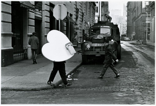 Black & white photo of a street in the 1960's. There's a person walking on the sidewalk, a lorry parked next to it. In front of the lorry there's a person crossing the street from right to left and another person from left to right. This latter person carries a very large white heart. 

The image source is: https://flic.kr/p/5y1X2s

The photo title is "Waverly Place" by "James Jowers" and it was shared by the George Eastman Museum on Flickr Commons. The photo is in the Public Domain.