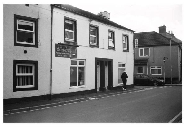 Black and white film photograph showing the front of a terraced street.  A sign affixed to the wall is for the Lotus Garden, Peking and Cantonese cuisine.