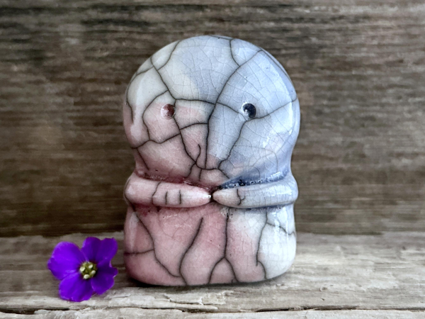 against a background of grey-brown driftwood, a kodama kami sculpture glazed in the pink, blue, and white of the transgender pride flag. it has a sweet and gentle face and two cute little arms. there's a violet flower next to it on the left.