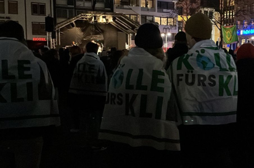 Bilck auf eine Bühne am Heumarkt nach Einbruch der Dunkelheit.

Im Vordergrund: Menschenm, die sich eine Kölle fürs Klima Fahne umgehängt haben.