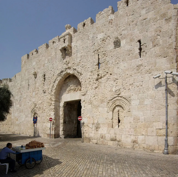 The image depicts a historic stone wall, likely part of a city fortification. There is a prominent archway in the center, which suggests it is a gate. In front of the wall, a person is seated at a small cart, possibly selling goods. The area appears to be a cobblestone street, and there are some road signs, including "no entry" signs. The sky is clear, indicating a sunny day.

Image Credits: Wikimedia / Berthold Werner / CC BY-SA 3.0