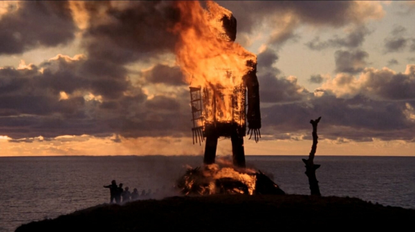 A burning wicker man next to the ocean. The skies are cloudy and it's sunset. People are gathered around holding hands and singing.