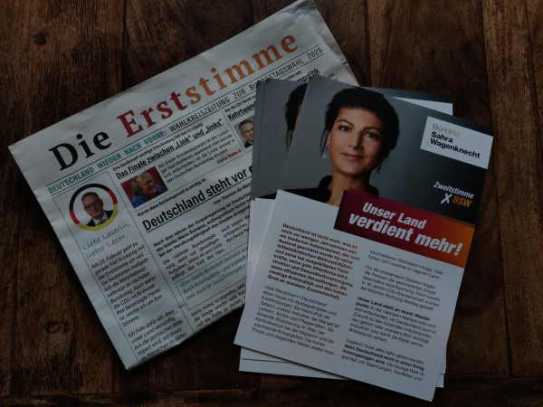 Zu sehen ist eine Zeitung mit dem Namen „Die Erststimme“ in den Deutschlandfarben Schwarz, Rot, Gold. Es handelt sich dabei um eine sog. Wahlkreiszeitung der cdU Leipzig zur Bundestagswahl 2025. Weiterhin zu sehen sind mehrere Flyer des BSW, das mit einem Foto von Sahra Wagenknecht um die Zweitstimme bei der Bundestagswahl wirbt.