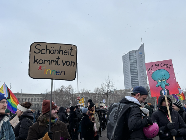 Foto von der Demo am Leuschnerplatz, im Hintergrund der Uniriese. Ein Demoschild mit der Aufschrift „Schönheit kommt von *innen“