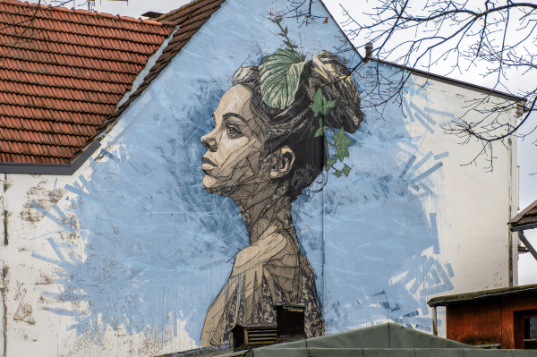 Das Bild zeigt ein Wandgemälde einer Frau mit Pflanzen im Haar, das an einer Hauswand angebracht ist.


Das Bild zeigt ein detailliertes, großformatiges Wandgemälde auf der Seite eines Gebäudes. Das Gemälde zeigt die stilisierte Darstellung einer Frau mit dem Kopf leicht nach oben geneigt. Ihr Gesicht ist im Profil zu sehen, wobei ihr Blick in die Ferne gerichtet ist.  Ihr Haar ist hochgesteckt und mit verschiedenen grünen Pflanzen und Blättern verziert, die wie eine Krone wirken. Der Stil des Wandgemäldes ist grafisch und detailliert, mit schraffierten Linien, die die Textur von Haut und Pflanzen darstellen. Die Farben sind hauptsächlich in gedeckten, natürlichen Tönen gehalten: Beige, Braun und verschiedene Blautöne für den Hintergrund. Der Hintergrund des Wandgemäldes scheint ein abstrakter blauer Farbverlauf zu sein, der an einen Himmel oder eine Landschaft erinnern könnte. Über dem Wandgemälde ist ein Stück des Gebäudes zu sehen, dessen Dach aus roten Ziegeln besteht und teilweise beschädigt zu sein scheint. Über dem Dach ragen trockene Äste eines Baumes hervor. Die Atmosphäre des Bildes ist ruhig und nachdenklich, mit einer gewissen Melancholie, die durch den Ausdruck der Frau und den Kontrast zwischen den lebendigen Pflanzen und dem etwas verwitterten Gebäude vermittelt wird.  Das Bild wirkt insgesamt künstlerisch und expressiv, möglicherweise mit dem Ziel, die Schönheit der Natur und die Verbindung zur Erde zu zeigen.
