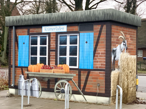 Ein bunt und freundlich bemaltes „Versorungshäuschen“ am Wochenmarkt. Mit täuschend echt wirkenden Sprossenfenster und blauen Fensterläden. Davor, auch gemalt, ein Handwagen mit Kürbissen und Gemüsen, eine Milchkanne. Rechts am Häuschen eine Ziege im Stroh. Krämmerladen steht über den Fenstern. 