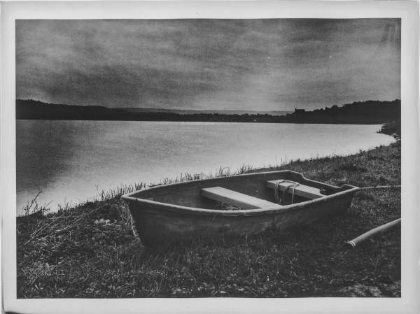 The image is a black and white photograph of a small wooden rowboat resting on the grassy shore of a calm lake. The boat appears slightly aged, with visible ropes inside. The water extends into the distance, reflecting a moody sky with a gradient of light and dark tones. In the background, a faint silhouette of a church or a similar structure can be seen on the horizon, surrounded by trees and distant hills. The overall atmosphere is serene and slightly melancholic.

The photograph is an optical print created in a darkroom using the lith print technique. This process is known for producing high-contrast images with soft, grainy textures and warm to cold tones, depending on the paper and developer used. The characteristic deep blacks and glowing highlights contribute to the dramatic and artistic feel of the scene.