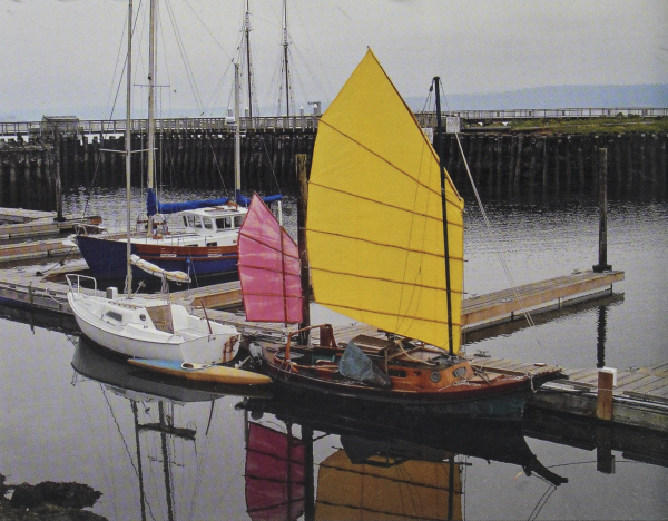 A color photo of a small two masted sailboat docked in a harbor. The ribbed mainsail is bright yellow and the other is candy pink.