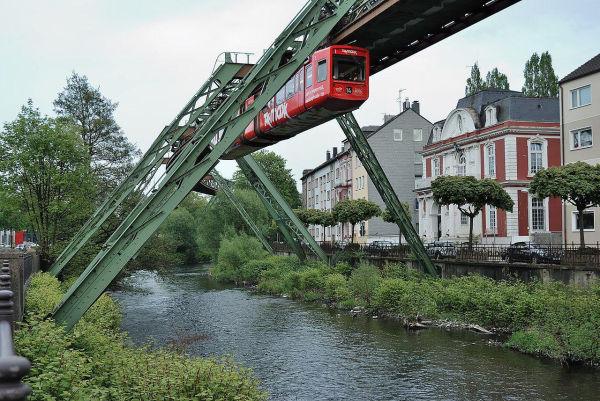 Ein Hochbahnwagen fährt auf einer schräg verlaufenden, grünen Schiene über einen Fluss. Im Hintergrund sind einige Gebäude und Bäume sichtbar, die sich am Ufer des Flusses befinden. Die Szenerie ist von einer grünen Umgebung umgeben.