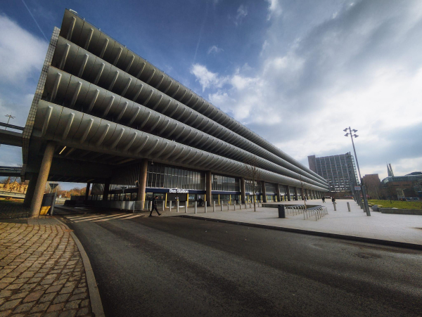 The image features Preston bus station, characterised by its modern architecture. The building has a prominent, expansive overhang supported by tall columns, creating a sheltered area for pedestrians below. The façade is sleek and features large glass windows, allowing natural light to illuminate the interior. In the foreground, a smooth, paved area is visible, with a few people walking and some bicycle racks. The sky above is partly cloudy, adding a dynamic element to the scene, while a multi-storey building can be seen in the background, contributing to the urban landscape.