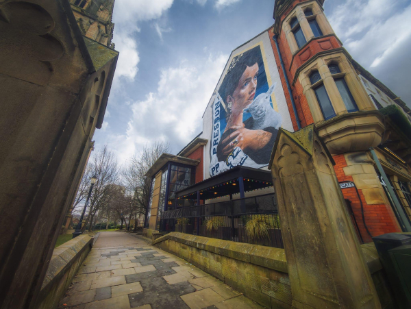 A vibrant mural on the side of a building in Preston, depicting a woman with intricate tattoos, cradling a white lamb. The artwork is framed by the brick and stone architecture of the building, which features large windows and ornate details. In the foreground, a cobbled pathway leads towards the mural, flanked by trees that are bare, suggesting early spring or late autumn. The atmosphere is lively, with string lights visible on the terrace below the mural, and the sky above is partly cloudy, adding depth to the scene.