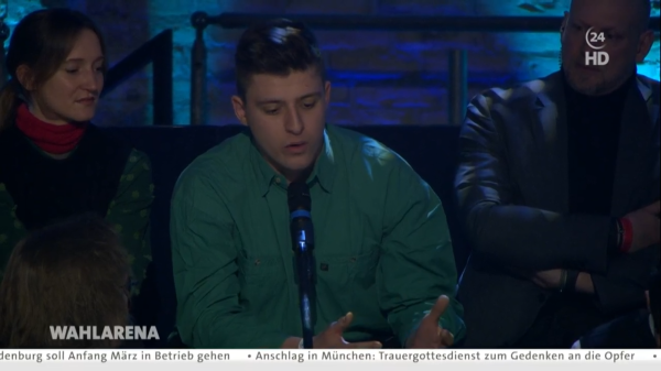 Screenshot der ARD Wahlarena vom 17.2.25. Auf dem Bild ein junger Mann in grüner Jacke mit einem Mikrofon und weitere junge Zuschauer:innen. Quelle:
https://www.tagesschau.de/newsticker/liveblog-bundestagswahl-montag-104.html#Uebertragung-auf-tagesschau24