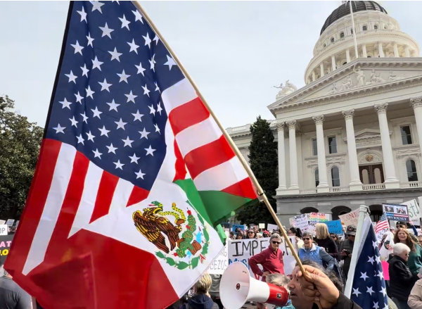 A protest is taking place in front of the California State Capitol. Flags of the United States and Mexico are prominently displayed. Participants hold signs and one person uses a megaphone, while a crowd gathers in the background.