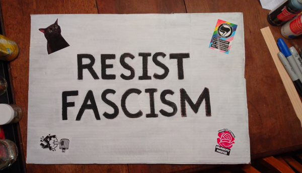 A sign with a white background and black letters. The sign reads: RESIST FASCISM
The sign also has some antifascist stickers, including "Resistance Cat" (an angry black cat).