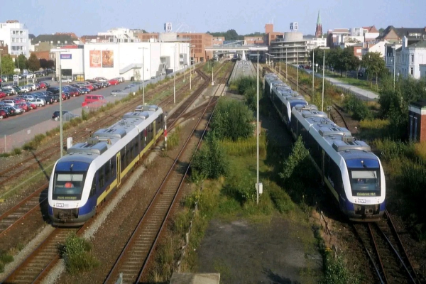 Zwei moderne Züge der Nordwestbahn, Baureihe VT 520, stehen auf einem zweigleisigen, geraden Gleis in einem urbanen Umfeld.
Linker Zug:
 * Silbernes Fahrzeug mit blau-gelber Aufschrift "Nordwestbahn" an der Seite.
 * Frontpartie mit großem, rechteckigem Fenster, zwei kleineren Fenstern darüber und einem mittigen, blau beleuchteten Zugzielanzeiger.
 * Seitliche Fensterfront mit mehreren kleinen, rechteckigen Fenstern.
 * Schwarzes Dach mit Aufschrift "GAAN".
Rechter Zug:
 * Gleiches Design wie der linke Zug, jedoch mit Fahrtzielanzeiger "Bremerhaven Hbf" unterhalb der Frontscheibe.
 * Frontpartie mit ähnlicher Fensteranordnung und mittiger Tür.
 * Seitliche Fensterfront mit gleichen, kleinen, rechteckigen Fenstern.
Weitere Details:
 * Beide Züge sind sauber und unbeschädigt.
 * Das Gleis verläuft gerade und eben.
 * Im Hintergrund sind weitere Gebäude und eine Brücke erkennbar, die jedoch unscharf sind.
 * Am linken Bildrand befindet sich ein Parkplatz mit mehreren Autos.
 * Der Himmel ist leicht bewölkt und hell.
 * Das Bild vermittelt einen Eindruck von Modernität und Funktionalität des öffentlichen Nahverkehrs.
Zusätzliche Informationen für Sehbehinderte:
 * Die Züge sind das Hauptmotiv des Bildes und nehmen einen Großteil des Raumes ein.
 * Die Perspektive ist leicht erhöht, wodurch die Züge und ihre Details gut sichtbar sind.
 * Die Farben sind dezent und natürlich.
 * Das Bild ist hell und freundlich.