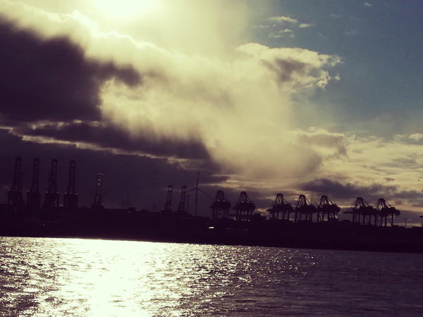 Blick aufs Wasser über den Fluss Elbe ans andere Ufer, der Sonne entgegen. Die strahlt in der linken oberen Ecke durch dunkle Wolken hervor. Die rechte Seite hat noch blauen Himmel. Im Hintergrund beginnt der Hamburger Hafen mit mehreren Kränen als Sillouetten vor sehr dunklen Wolken die heranziehen. 