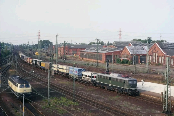  Eine dunkelgrüne Elektrolokomotive der Baureihe 140 mit auffällig silbergrauem Dach zieht am 13. August 1990 einen Güterzug des kombinierten Ladungsverkehrs durch den Bahnhof Hamburg-Harburg in südlicher Richtung. Der Zug besteht aus verschiedenen Containern und Wechselbrücken, darunter Werbeaufschriften wie „DEA“. Im Hintergrund sind die roten Backsteinhallen des ehemaligen Güterwagen-Ausbesserungswerks zu sehen, das später einem Baumarkt weichen sollte. Links im Bild steht eine beige-blaue Diesellokomotive der Baureihe 218 auf einem Abstellgleis. Das Bahngelände ist von zahlreichen Gleisen, Oberleitungen und Signalanlagen geprägt.
