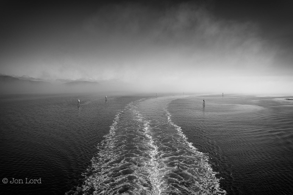 A High Contrast Black And White Seascape Photo In Landscape Format. The Lower Half Of The Image Is Taken Up Mostly By A Body Of Jet Black Sea Water. In The Bottom Centre And Rising Upwards Towards The Middle Before Angling To The Right Is The White Wake Of A Passing Ship. Either Side Of The Ship's Wake Are Channel Markers Made Up Of A Tripod Of Long Poles With A Circular Marker And Light On Top. Above The Water And To The Left Is The Distant Rising Terrain Of A Range Of Hills Or Small Mountains. To The Right Are A Few Small Skerries, Just Visible Above The Water. Above The Water Is An Expanse Of Sea Fog Illuminated By The Rising Sun. Above The Fog A Jet Black Clear And Cloudless Sky.
The Location Is The Risøyrenna, A Dredged Deep Water Channel Opened In 1922 Allowing Ships To Avoid The Open Waters Rounding Andøya Island And The Perilous Norwegian Sea (2018).