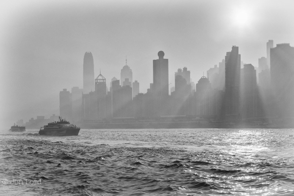 A Black And White Hazy Seascape / Cityscape Dawn Photo In Landscape Format. In The Foreground Is An Expanse Of Choppy Harbour Water That Extends Up One Third Of The Image. In The Lower Left Corner Are Two Small Ferries. The First And Closest Is A Fast Ferry (SeaCat) Underway From Left To Right. The Further, A Standard Ferry Is Underway From Right To Left And Approaching The Left Margin. Above The One Third Part Of The Photo Is A High-Rise Cityscape With The Tops Of The Buildings Approaching The Three Quarter Upwards Mark. In The Upper Right Corner Is The Rising Morning Sun Just Above The Buildings. The Sun Is Illuminating The Haze Or Thin Mist But The Buildings Are Causing Shadows Resulting In The Appearance Of Sun Beams Shining Out From Between The Buildings. 
The Location Is Victoria Harbour, 維多利亞港, And The Cityscape Is Hong Kong Island, 香港, (2018)