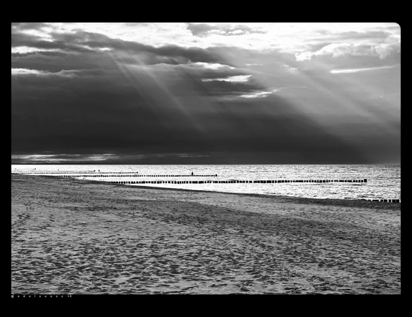 Schwarzweißfotografie: Blick seitlich über den weiten Sandstrand an der Ostseeküste am Abend, unzählige Spuren und Unebenheiten im Sand, im Hintergrund die anlandende Ostsee, hell durch die restliche Abendsonne spiegelnd, mehrere Buhnenreihen in Strandnähe, darüber dunkler Himmel mit hellen Lichtstrahlen, die sich kontrastreich durch die Wolken brechen, eingefasst ist das Bild in einen breiten schwarzen Rahmen 