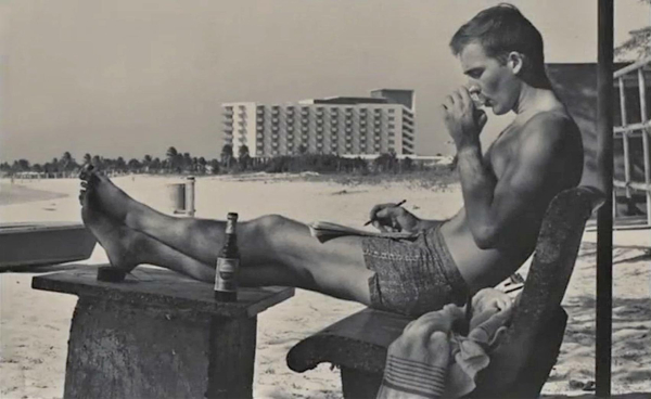 The author in profile seated on a bench in swimming trunks, his legs propped up on a table, on a beach in Puerto Rico. A large beachfront hotel is visible in the distant background. The black and white  photo appears on the cover of The Rum Diary (1959).