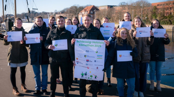 Ministerpräsident Günther und Vertreterinnen und Vertreter der teilnehmenden Kreise stehen im Sonnenschein an der Kieler Förde. Sie halten Plakate in den Händen, die auf den Klimathon 2025 hinweisen. 