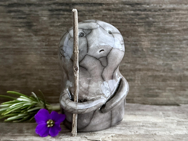 against a background of grey-brown driftwood, a raku ceramic warrior sculpture. it's glazed in a bottom-to-top gradient of darker to lighter beige-brown, and it carries an autumn-sage staff in its right arm. its face is desperately worry but also determined. 
next to it on the left are a sprig of rosemary and a deep purple flower.