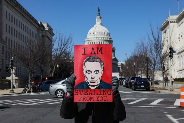 A protestor holds up a Musk "I am stealing from you" sign.