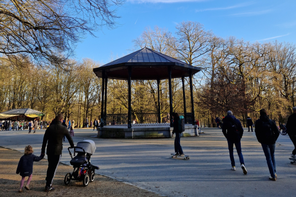 A bandstand in a park under blue skies and trees. People can be seen enjoying the open space. 