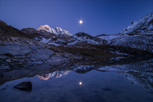 Photo d'un paysage de montagne au crépuscule.
Des montagnes saupoudrées de neige se reflètent nettement dans un lac. Un sommet plus éloigné est encore éclairé par le soleil, le reste de l'image est plongé dans l'ombre.
Dans le ciel, la lune brille et semble prendre une forme étoilée, elle se reflète aussi dans le lac. On distingue difficilement une ou deux étoiles dans le ciel.
-
Photo of a mountain landscape at dusk.
Snow-capped mountains are clearly reflected in a lake. A more distant peak is still illuminated by the sun, while the rest of the picture is plunged into shadow.
In the sky, the moon shines brightly and seems to take on a star-like form, also reflected in the lake. One or two stars are hard to make out in the sky.