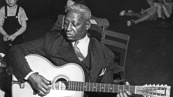 Black and white photo of Lead Belly. He’s sitting while playing an acoustic guitar. He’s wearing a suit. Some children are sitting and listening in the background.