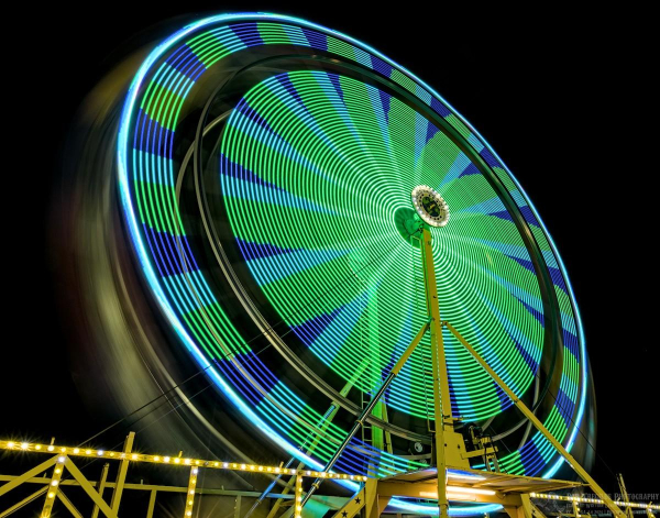 A long exposure of a spinning Ferris wheel. Leaving the camera shutter open causes the lights to make all kinds of interesting patterns. This one has spokes radiating out from the center of green, light blue, and dark blue. There's a wide black line near the out edge where the lights were off.